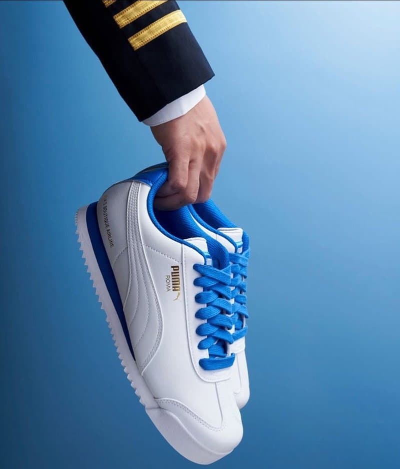 Zapatilla Puma Roma blanca y azul sostenida por una mano con manga de piloto y galones dorados