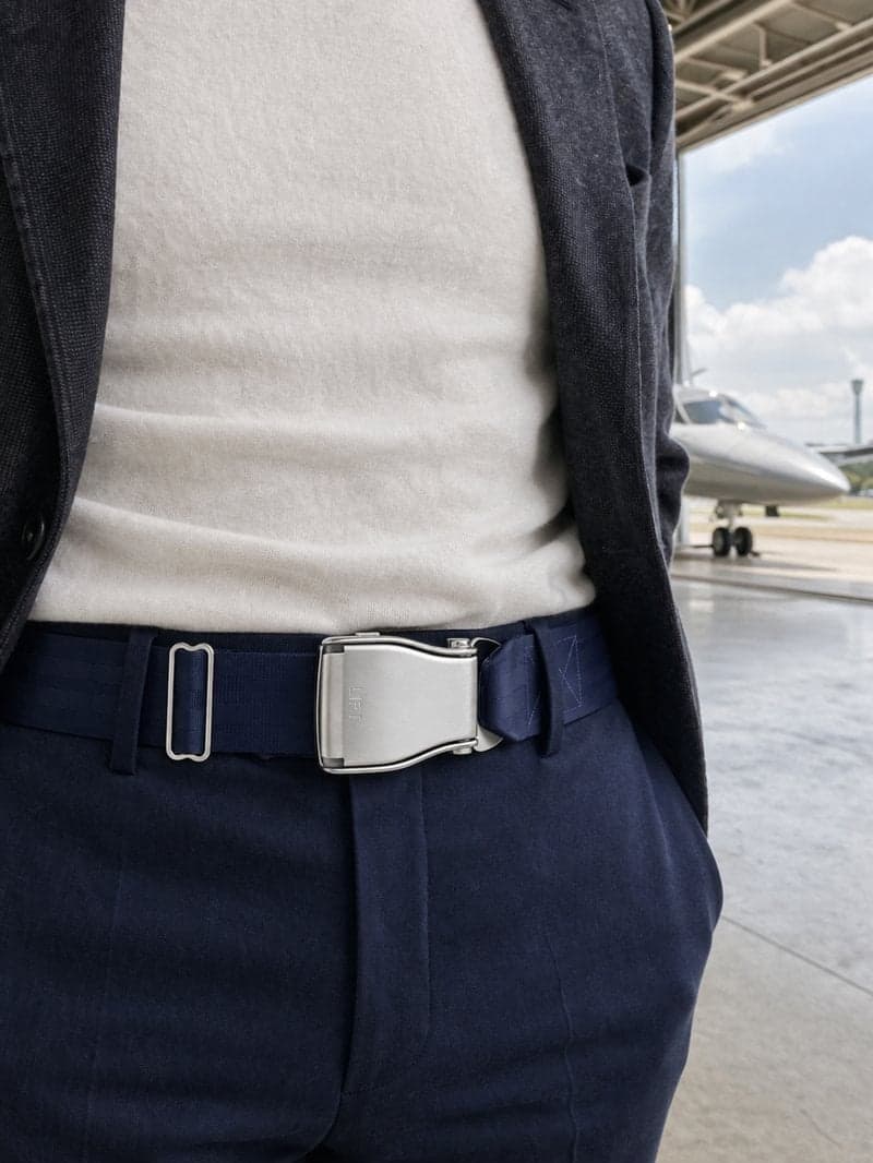 Ceinture d'avion TRANSATLANTIC bleu marine portée avec un pantalon bleu marine, un pull crème et un blazer anthracite, dans un hangar