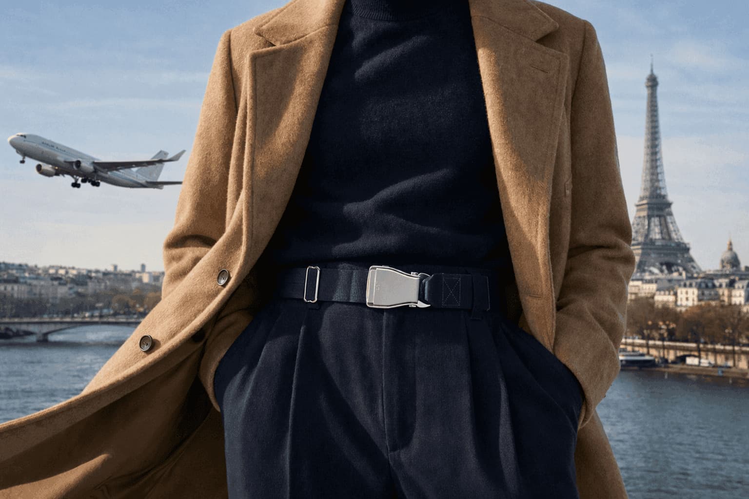 A man in a camel coat wearing the Fly-Belts Transatlantic navy belt in front of the Seine, the Eiffel Tower and a departing aircraft — Paris, Est. 2012.